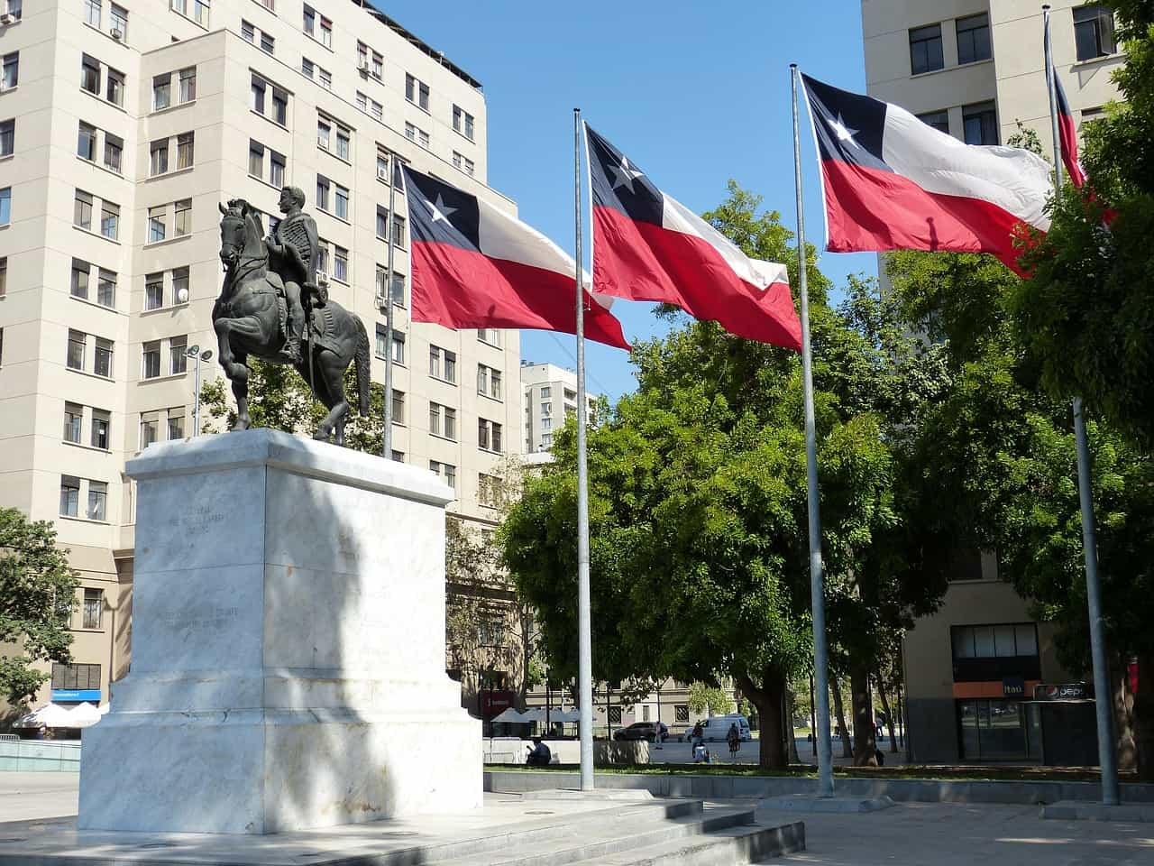 Plaza en Santiago de Chile, con un monumento de hombre a caballo y tres banderas del país ondeando desde sus respectivos mástiles.