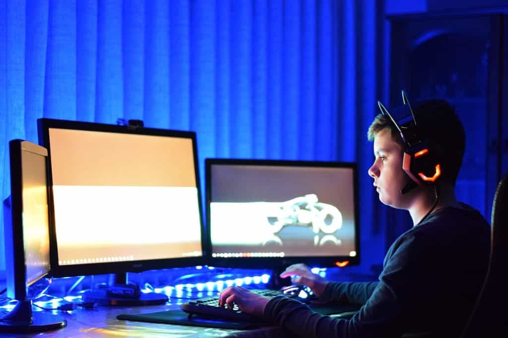 Niño con auriculares enormes frente a tres monitores en habitación poco iluminada.