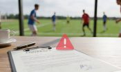 Contrato sobre una mesa con un icono de alerta, y un partido de fútbol desenfocado al fondo.