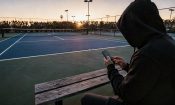 Persona con capucha usando un móvil junto a una pista de tenis al atardecer.