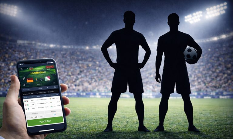 Teléfono móvil con aplicación de apuestas deportivas frente a dos futbolistas en estadio.