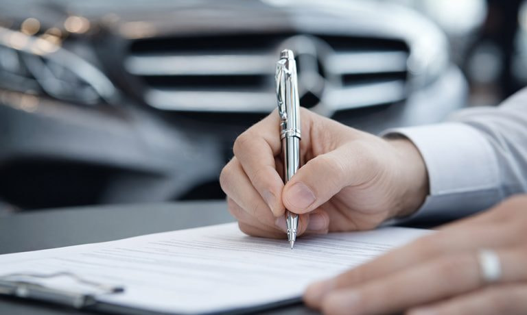 Mano firmando un documento con un coche desenfocado al fondo.
