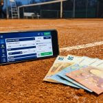 Teléfono móvil con apuestas de tenis junto a billetes en una pista de tierra batida