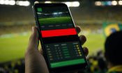 Mano sostiene un móvil con una app de apuestas y un aviso rojo en un estadio con bandera de Brasil.