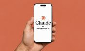 Mano sosteniendo un móvil con la pantalla de inicio de la app de Claude, de Anthropic, sobre fondo naranja.