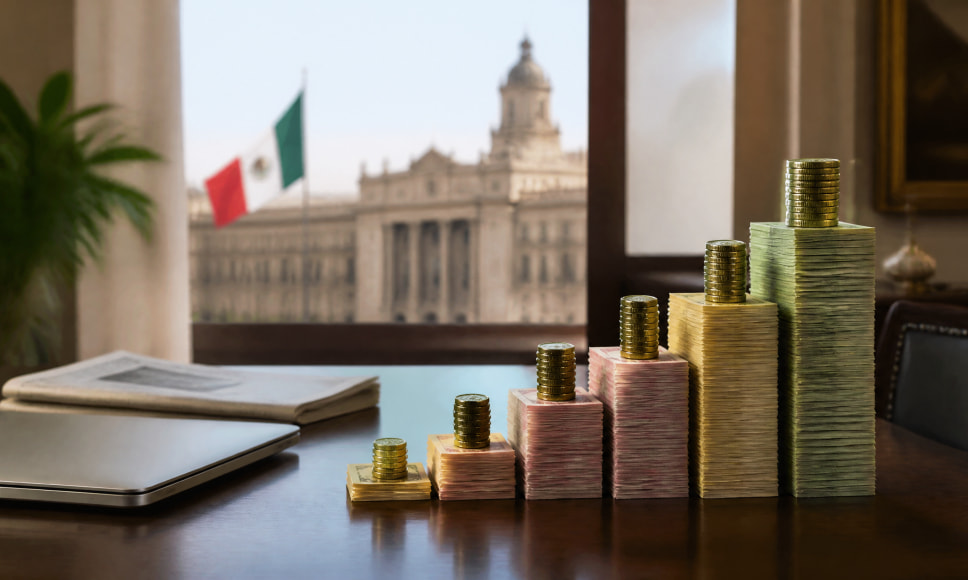 Pilas crecientes de billetes y monedas sobre un escritorio con la bandera de México y un edificio gubernamental al fondo.