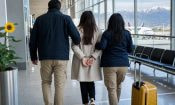 Mujer esposada escoltada por agentes en un aeropuerto con montañas de fondo y una maleta amarilla.